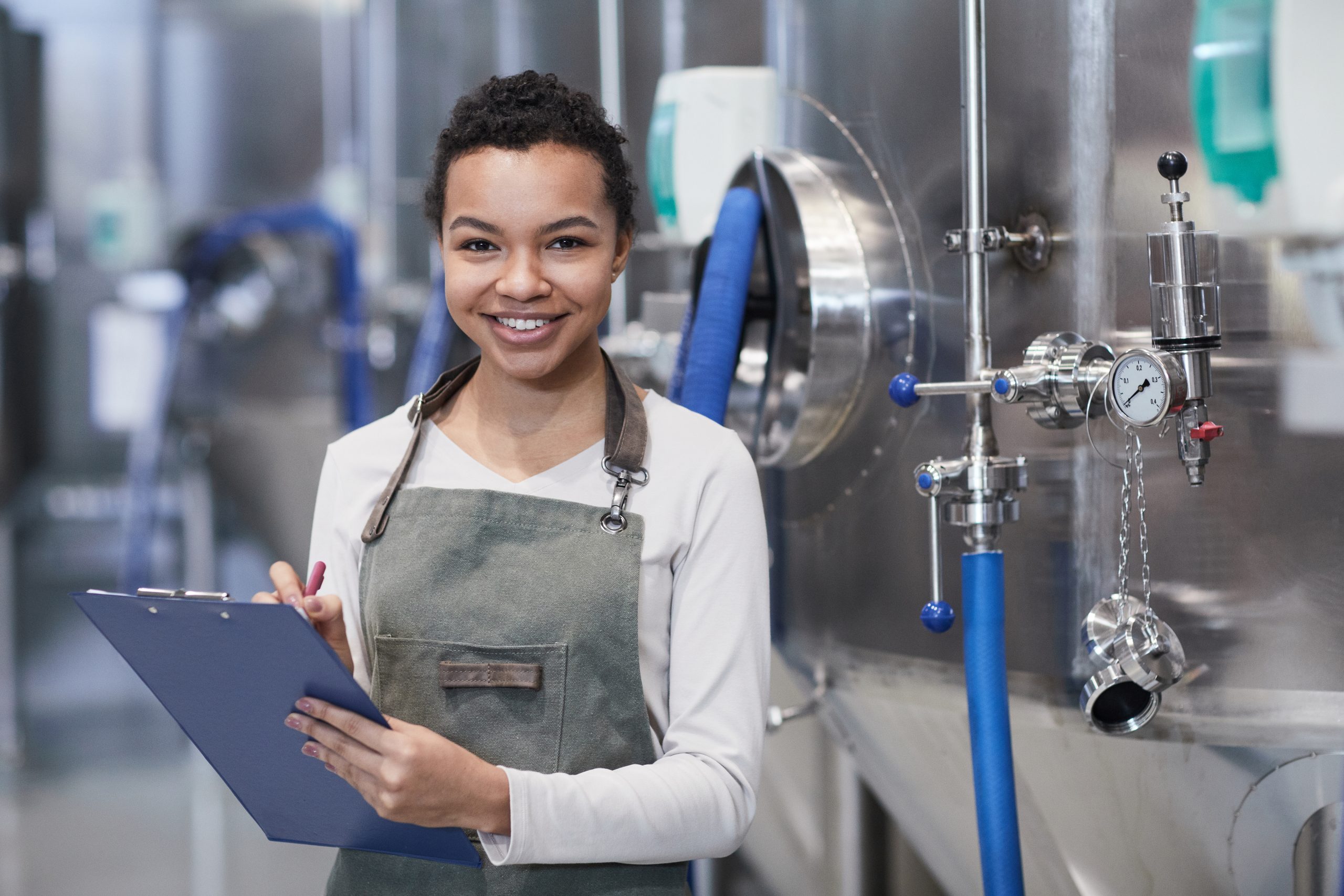 Waist up portrait of young African-American woman enjoying work at industrial factory and smiling at camera holding clipboard, copy space